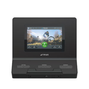 A black fitness console with a color touchscreen displaying workout options, outdoor scenery, and controls. The brand name TRUE is visible below the screen. The console also features caution and warning labels at the bottom.